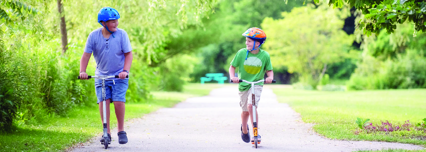 Two young boys riding scooters down a paved path in a park
