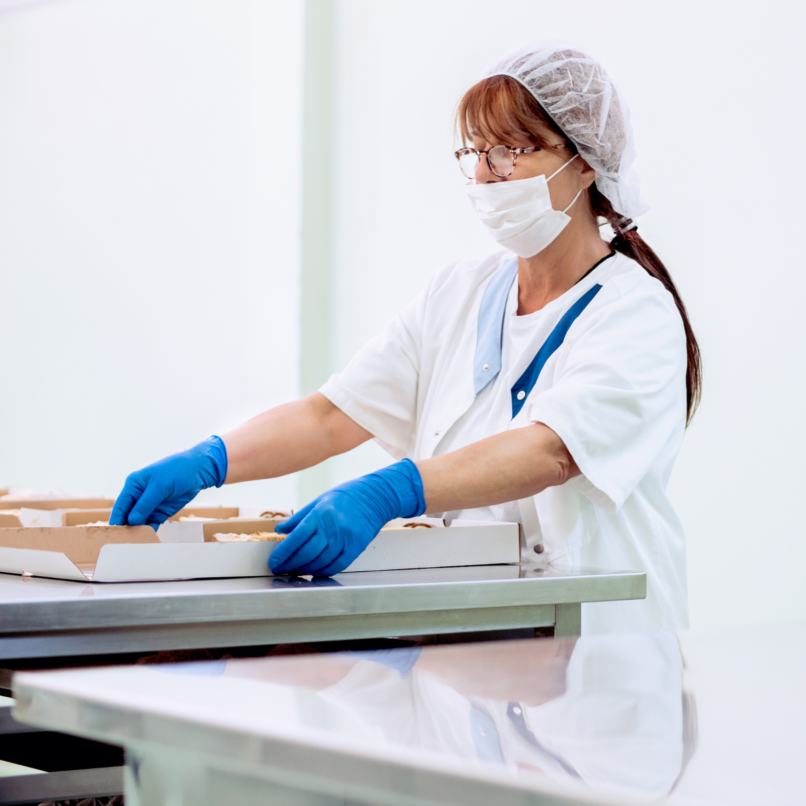 Food service worker wearing gloves, mask, and hairnet packaging food in a commercial kitchen