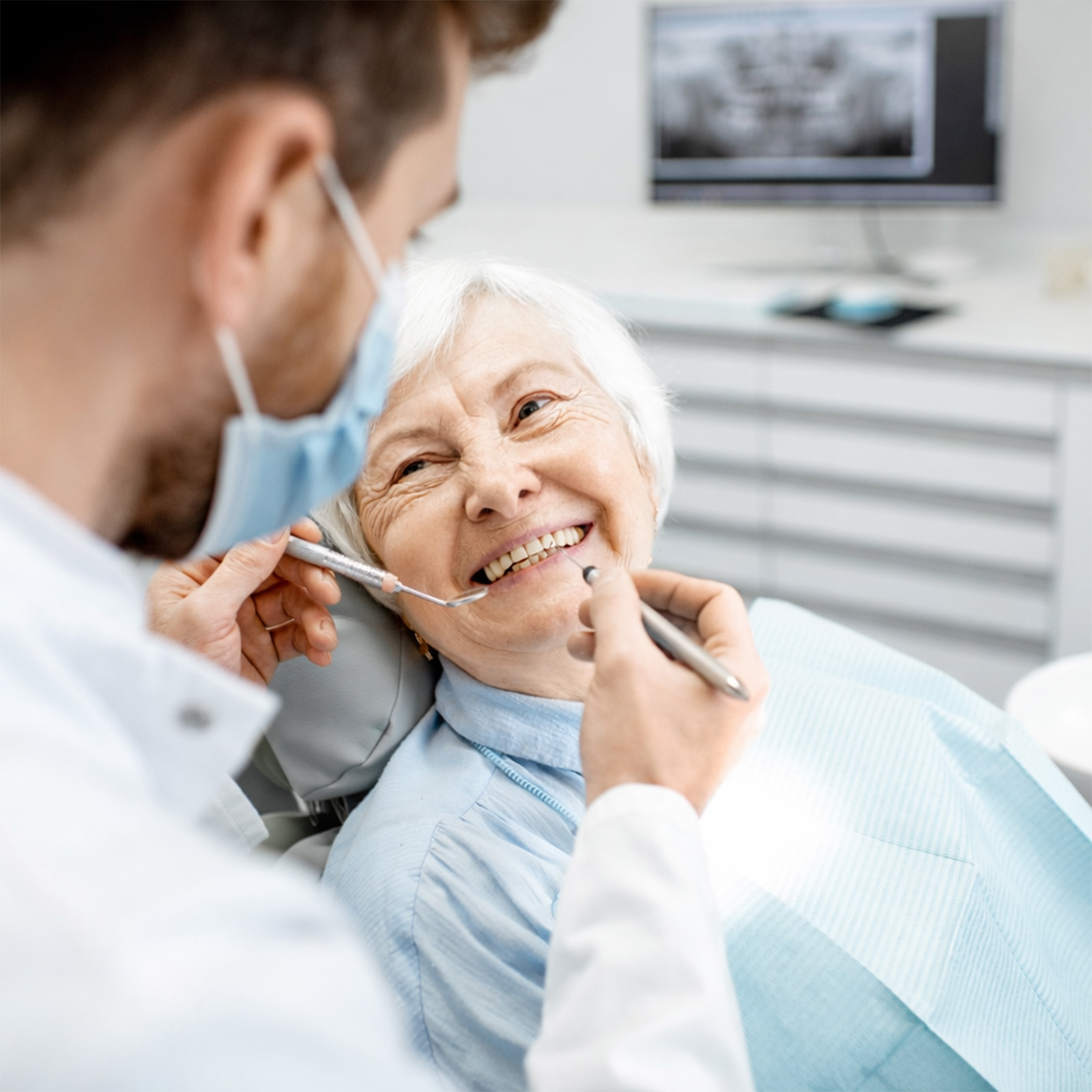 A dentist wearing a face mask examines an elderly woman’s teeth while she smiles in a dental chair.