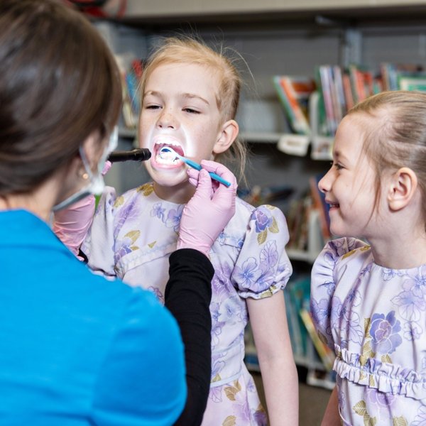 Dental hygienist examining a child’s teeth while another child watches and smiles