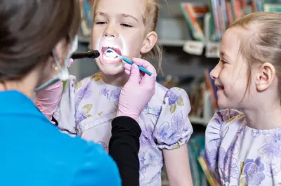 Dental hygienist examining a child’s teeth while another child watches and smiles