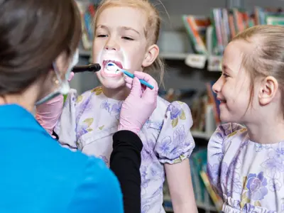 Dental hygienist examining a child’s teeth while another child watches and smiles